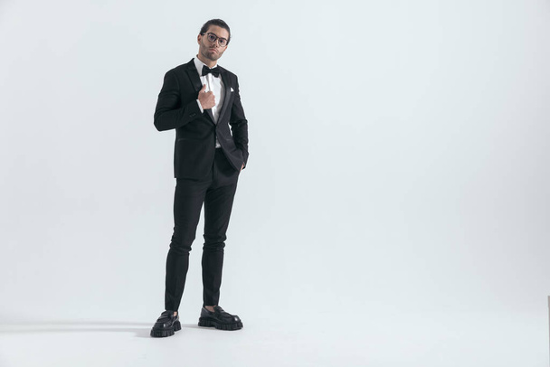 sexy giovane uomo con gli occhiali regolazione smoking nero e posa con mano in tasche mentre in piedi su sfondo grigio - Foto, immagini