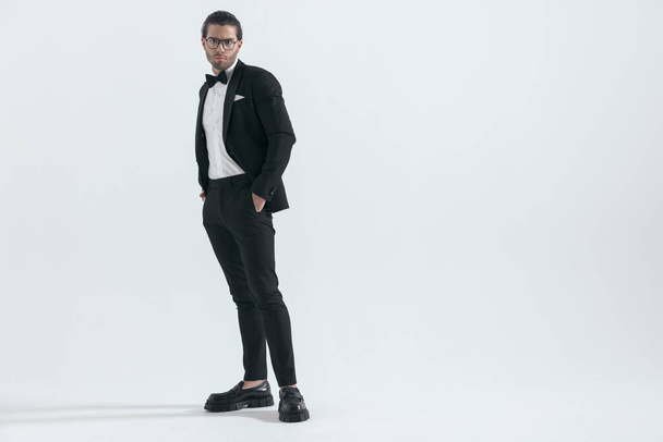 young fashion man in black tuxedo holding hands in pockets and standing in front of grey background - Photo, Image