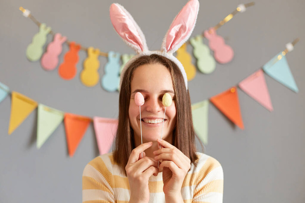 Indoor shot of smiling funny woman wearing rabbit ears hiding her eye with Easter cake pops isolated on gray decorated background, having fun during holiday celebration - Фото, изображение