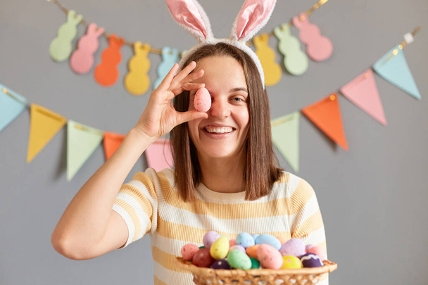 Indoor shot of extremely happy optimistic woman wearing rabbit ears holding Easter eggs in wicker basket, covering her eye with egg, smiling to camera having fun, isolated on gray decorated background - Fotó, kép
