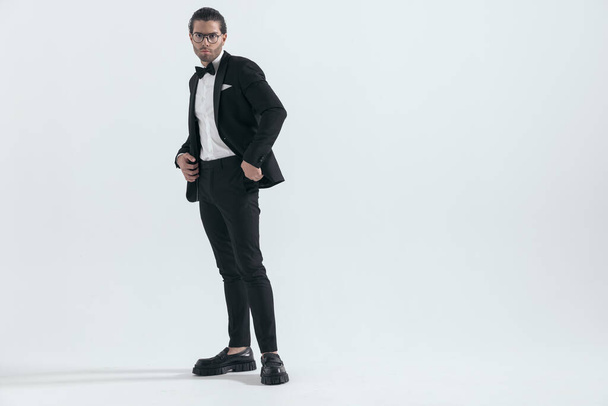 elegant young groom in tuxedo holding hand in pockets and standing in front of grey background, full body, - Foto, Imagen