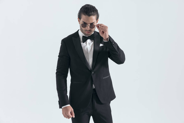 elegante novio en esmoquin negro ajustando gafas de sol y posando con confianza delante de fondo gris - Foto, imagen