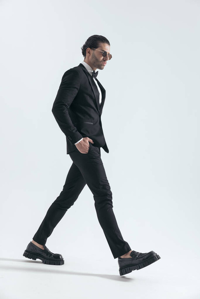 κομψό νεαρό επιχειρηματία με μαύρο σμόκιν περπατώντας και κοιτάζοντας προς τα πλάγια κρατώντας τα χέρια σε τσέπες σε γκρι φόντο - Φωτογραφία, εικόνα