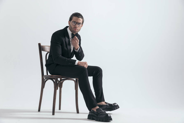 serious elegant man in black tuxedo frowning and fixing bowtie while sitting on wooden chair on grey background - Photo, Image