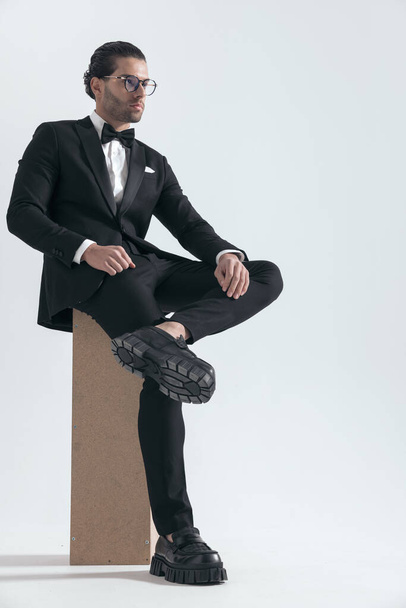 atraente homem elegante olhando para o lado enquanto sentado com o tornozelo no joelho na frente de fundo cinza - Foto, Imagem