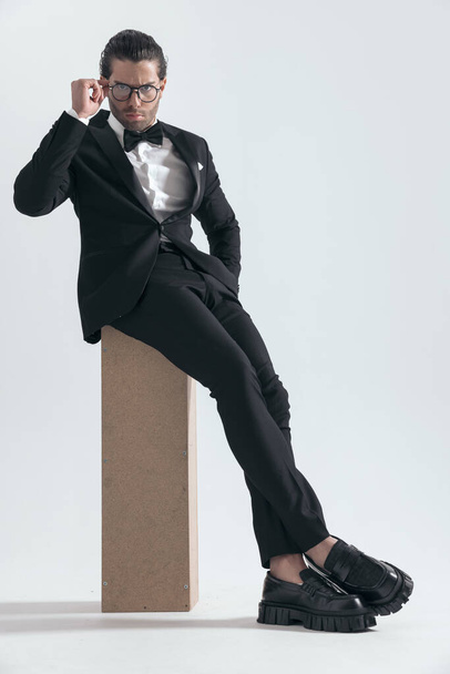 elegant young groom in black tuxedo fixing glasses and sitting with hand in pocket in front of grey background - Photo, Image