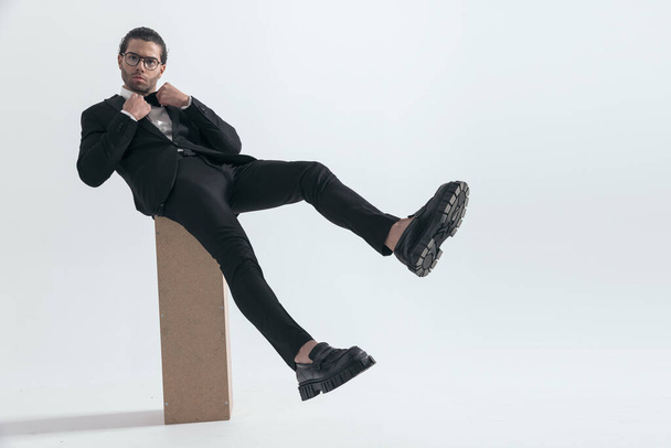 jóképű hűvös férfi szemüveggel állító csokornyakkendő, miközben ül lábak előtt szürke háttér - Fotó, kép