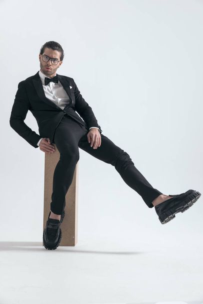 sexy muž v černém smokingu s brýlemi sedí s nohou ve vzduchu a pózuje chladným způsobem na šedém pozadí - Fotografie, Obrázek