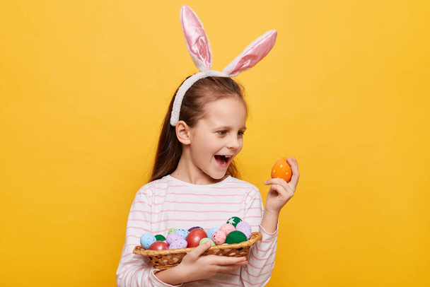 Indoor shot van extreem gelukkig klein meisje draagt konijnenoren met paaseieren in rieten mand, winnende Paasjacht, schreeuwend van opwinding en geluk geïsoleerd op gele achtergrond - Foto, afbeelding