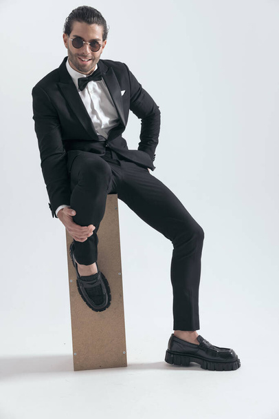 sexy junger Mann sitzt mit der Hand in der Tasche, hält das Bein und lächelt, während er auf grauem Hintergrund sitzt - Foto, Bild