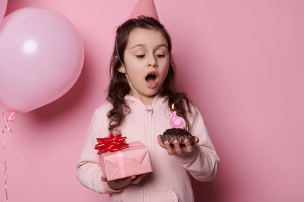 Schattig 5 jaar oud blank kind, mooie verjaardag meisje in feestelijke roze feestmuts, met een geschenkdoos met een gelukkig cadeau en blaas de kaars op een feestelijke taart, geïsoleerd op roze achtergrond - Foto, afbeelding