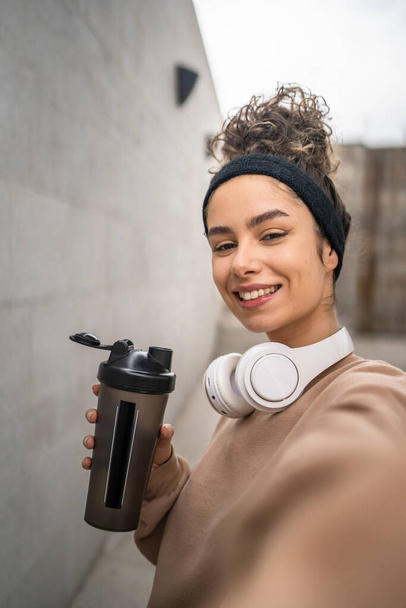 One young woman with headphones prepare for training hold supplement shaker bottle to drink water stand outdoor beautiful sporty caucasian female selfie healthy lifestyle UGC User generated content - Photo, Image