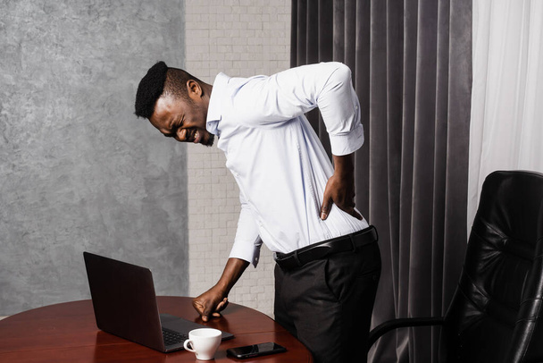 African manholds his back and feels pain in his back and neck after working for long hours with laptop online in office. Cervical spine osteochondrosis is radicular syndromes of african american man - Fotografie, Obrázek