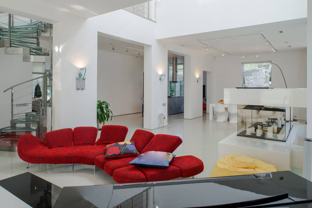 Modernes Interieur eines luxuriösen Privathauses. Geräumiges Wohnzimmer mit rotem Sofa, weißem Tisch. Wendeltreppe aus Metall und Glas. - Foto, Bild