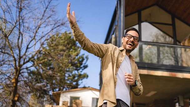 Um homem caucasiano adulto macho stand ao ar livre na frente do anfitrião casa pequena moderna ou vendedor com mãos gesto convidar em convite gestual sorriso feliz no espaço de cópia dia ensolarado - Foto, Imagem
