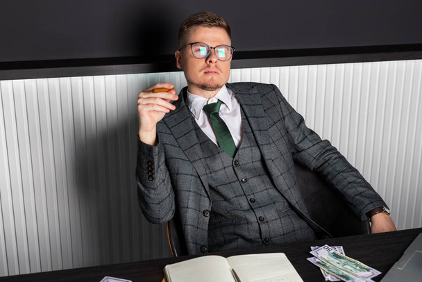Mladý vážný muž v šedém obleku a brýlích sedí za stolem obklopený penězi nebo penězi a kouří doutník. Podnikatel v práci. - Fotografie, Obrázek
