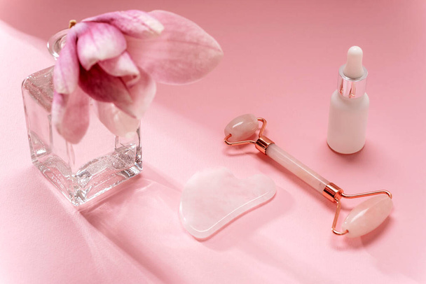 Rose quartz crystal facial roller and gua sha scraper, face serum and magnolia flower in vase on pink background. Facial massage kit for lifting therapy . Skin care anti-aging tools. Top view. - Fotó, kép