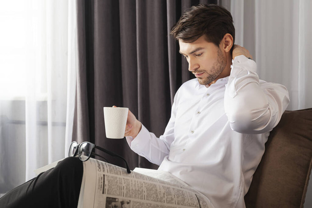 Ένας όμορφος νέος με ρούχα που κάθεται σε έναν καναπέ και διαβάζει εφημερίδα με ένα φλιτζάνι καφέ στο χέρι του σε βιομηχανικό στυλ δωμάτιο.. - Φωτογραφία, εικόνα
