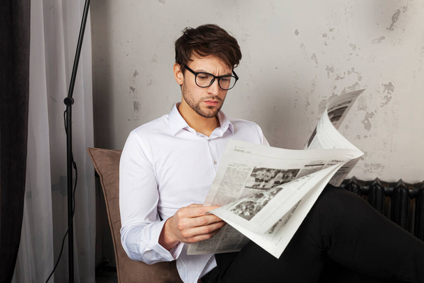 Elbiseli yakışıklı genç adam koltukta oturuyor ve endüstriyel tarzda gazete okuyor.. - Fotoğraf, Görsel