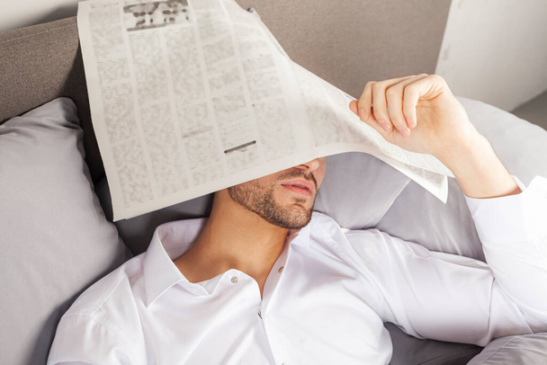 Ein hübscher junger Mann in weißem Hemd schlief auf dem Bett ein, während er eine Zeitung las. Gesicht mit einer Zeitung bedeckt. - Foto, Bild