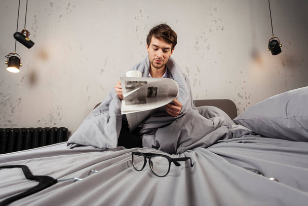 Un joven sonriente está sentado en una cama, envuelto en una manta, y leyendo buenas noticias de un periódico con una taza de café en la mano. - Foto, imagen