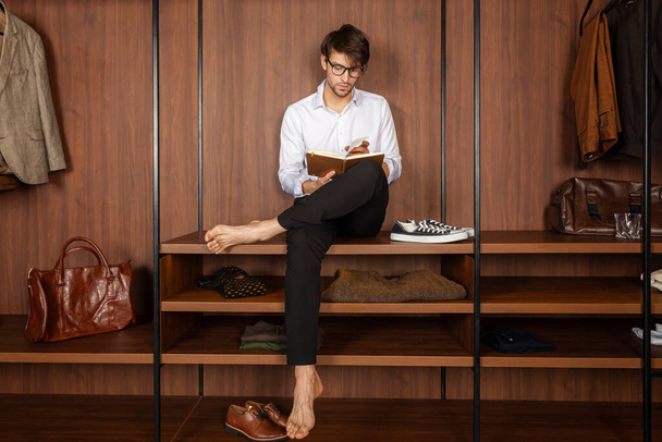 Beau jeune homme en vêtements habillés assis sur une étagère à vêtements et lisant un livre. Vue de face. - Photo, image