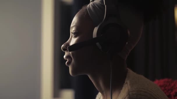 Afrykańska kobieta grać w grę za pomocą joystick siedzi w ciemnym salonie .Close-up dziewczyna nosić słuchawki i trzymając gamepad podczas gry online w domu.Czarna pani cieszyć się gry wideo mówienie - Materiał filmowy, wideo