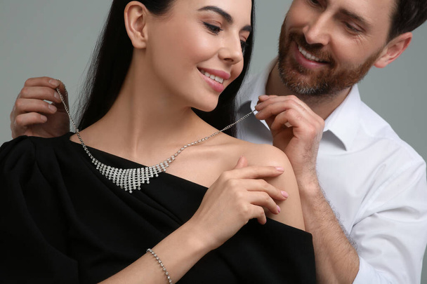 Man putting elegant necklace on beautiful woman against grey background, closeup - Foto, afbeelding