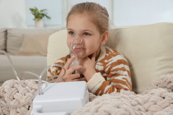 Little girl using nebulizer for inhalation in armchair at home - Foto, afbeelding