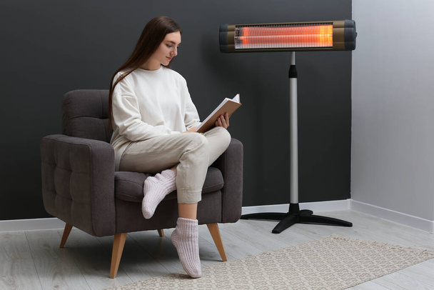 Young woman reading book on armchair near electric infrared heater at home. Space for text - Photo, image