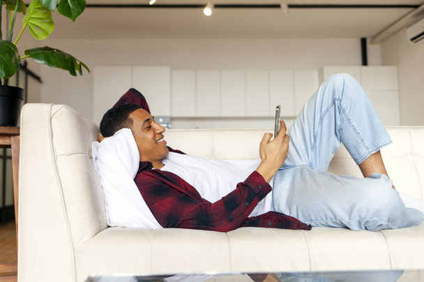young african american man lies on comfortable sofa and uses smartphone, guy holds and looks at the phone at home and smiles, man is resting and holding mobile phone - Фото, изображение