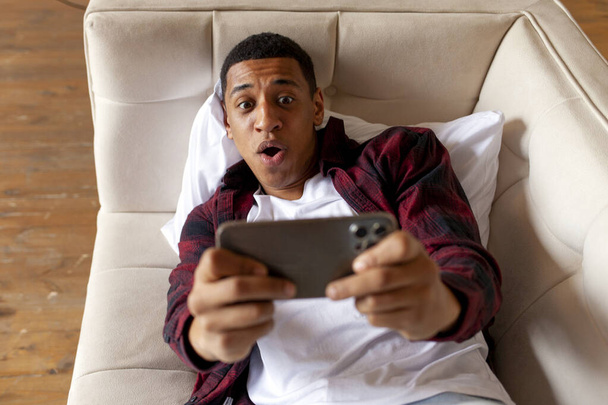молодий здивований афроамериканський чоловік лежить на зручному дивані і грає в відеоігри на смартфоні, шокований хлопець геймер грає в мобільні ігри вдома
 - Фото, зображення