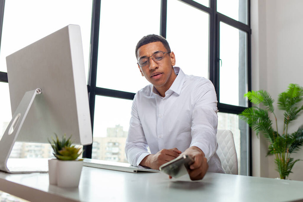 офисный работник за столом в офисе сидит рядом с компьютером и проверяет сообщение на смартфоне, африканский американский бизнесмен в офисе смотрит на телефон - Фото, изображение
