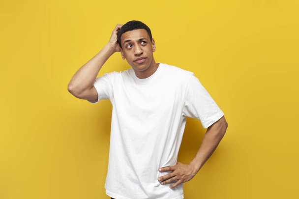 młody, zakłopotany Afrykański Amerykanin w białej koszulce szkicuje głowę dłonią na żółtym, odizolowanym tle, zdezorientowany człowiek myśli o problemie - Zdjęcie, obraz