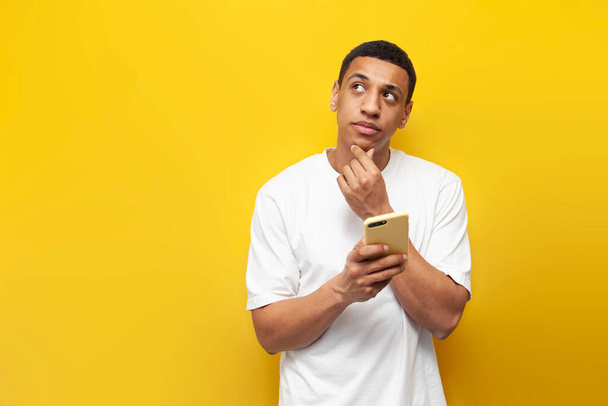 mladý zamyšlený africký Američan v bílém tričku používá smartphone a sny na žlutém izolovaném pozadí, přemýšlí muž a dívá se na kopírovací prostor s telefonem - Fotografie, Obrázek