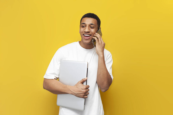 young african american guy in white t-shirt holds laptop and talks on the phone on yellow isolated background, man with modern gadgets communicates on smartphone - Фото, изображение
