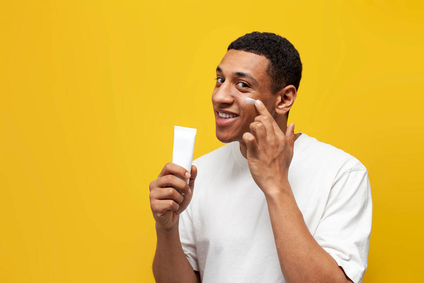 joven afroamericano chico en blanco camiseta muestra tubo de crema en amarillo aislado fondo, hombre anuncia cosméticos cara - Foto, Imagen