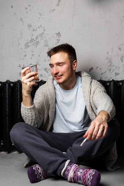 Ο πολύ μεθυσμένος νεαρός κάθεται στο πάτωμα δίπλα σε μια θερμάστρα με τσιγάρο και ένα ποτήρι αλκοόλ. μεθυσμένο πρόσωπο. - Φωτογραφία, εικόνα