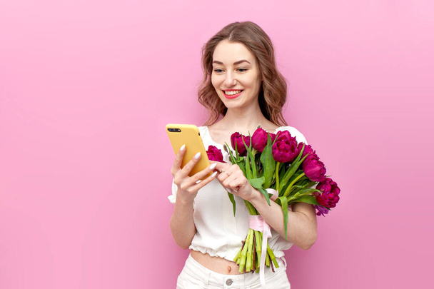 young attractive woman in white festive clothes holding bouquet of pink tulips and using smartphone on isolated background, girl with flowers looking on phone, concept of spring and women's holiday - Photo, image