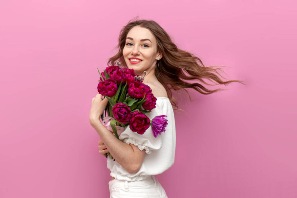 ピンクのチューリップの花束を持っている白いお祝いの服の若い魅力的な女性と孤立した背景に渦巻く、女の子は花と彼女の髪で移動し、春と女性の休日の概念を飛んでいる - 写真・画像