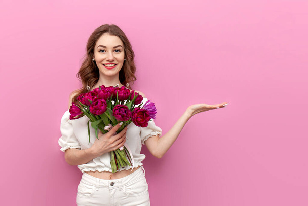 young woman in white clothes holding bouquet of pink tulips and pointing at copy space on isolated background, girl with flowers advertises place for text, concept of spring and women's holiday - Photo, Image