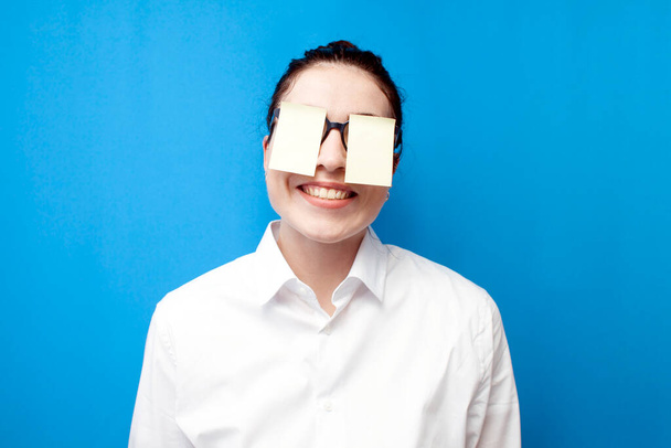 cheerful girl manager in white shirt with blank papers in front of her eyes smiles on blue background, woman office worker in glasses on colored background - Foto, immagini