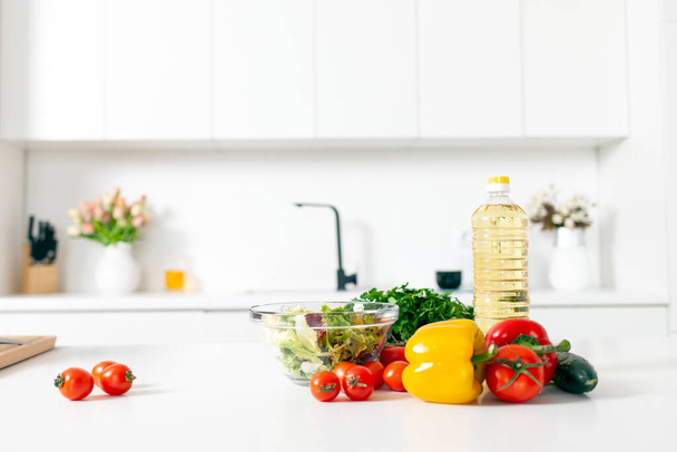 składniki sałatki i produktów leżą na stole w białej nowoczesnej kuchni, zieleni, pomidory, papryka, butelka oleju w pustym świetle kuchnia, zdrowe wegetariańskie jedzenie - Zdjęcie, obraz