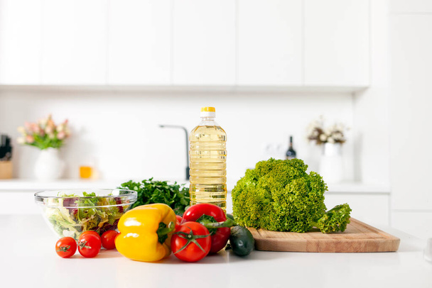 ingredientes para ensalada y productos se encuentran sobre la mesa en la cocina moderna blanca, verduras, tomates, pimientos, botella de aceite en la cocina vacía luz, comida vegetariana saludable - Foto, imagen