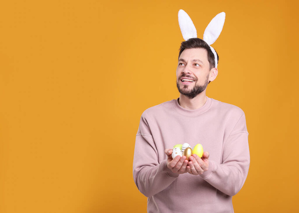 Happy man in bunny ears headband holding painted Easter eggs on orange background. Space for text - Fotoğraf, Görsel