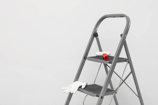 Metallic folding ladder, paint roller brush and rubber gloves near light grey wall, space for text - Fotoğraf, Görsel