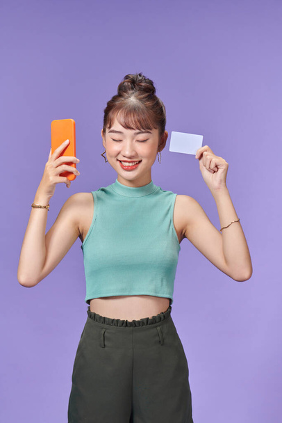 Akıllı telefon ve kredi kartını mor arka planda tutarken gülümseyen iyimser kadın - Fotoğraf, Görsel