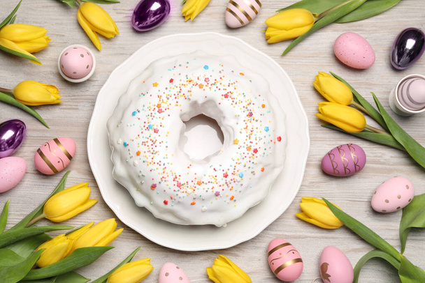 Easter cake with sprinkles, painted eggs and tulips on white wooden table, flat lay - Photo, Image