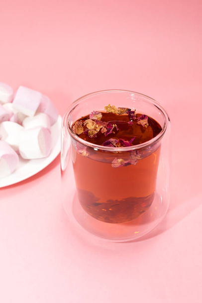 Healthy herbal rose petals, buds tea in double walled glass mug, cup. Pink white marshmallow plate. Pink background. Desiccated colorful flower petals herbal drink, beverage. Vertical, copy space. - Фото, изображение