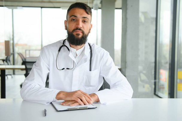Young 30-aged handsome pleasant Arabic male doctor in white coat, posing at camera indoors - Photo, image